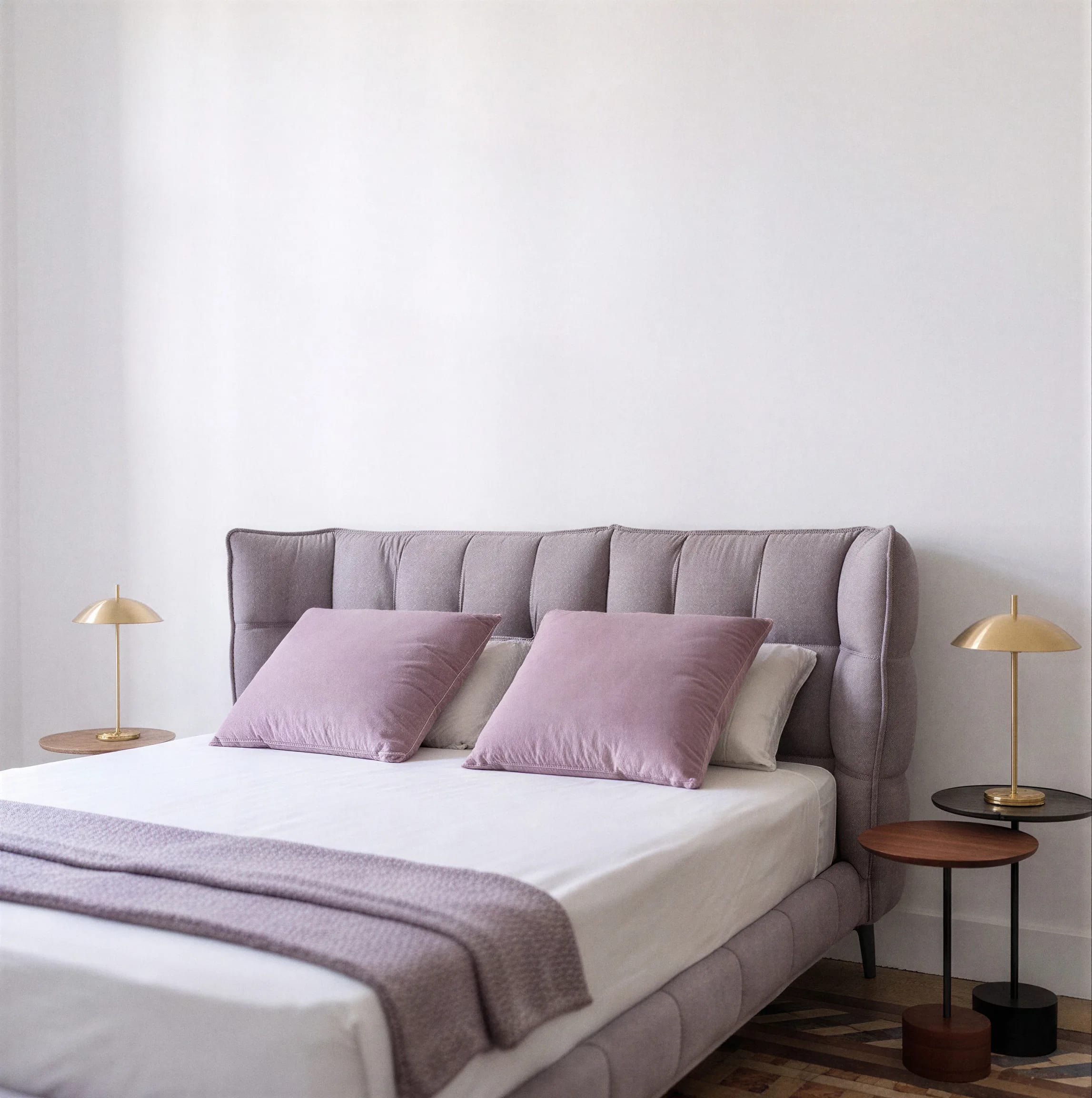 Cama tapizada con cojines lila y lámparas de latón sobre mesillas redondas